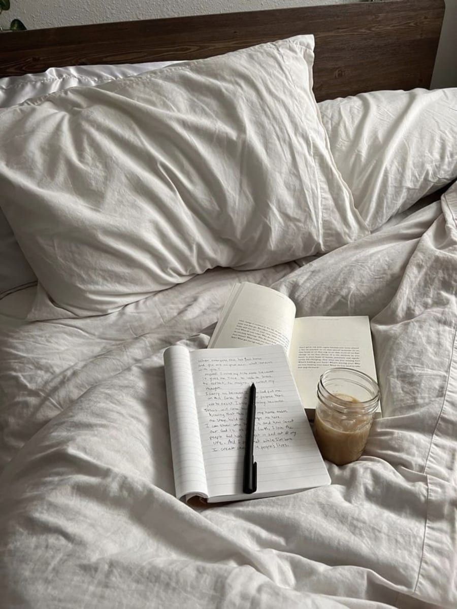 An open journal, pen and book on an unmade bed beside an iced coffee